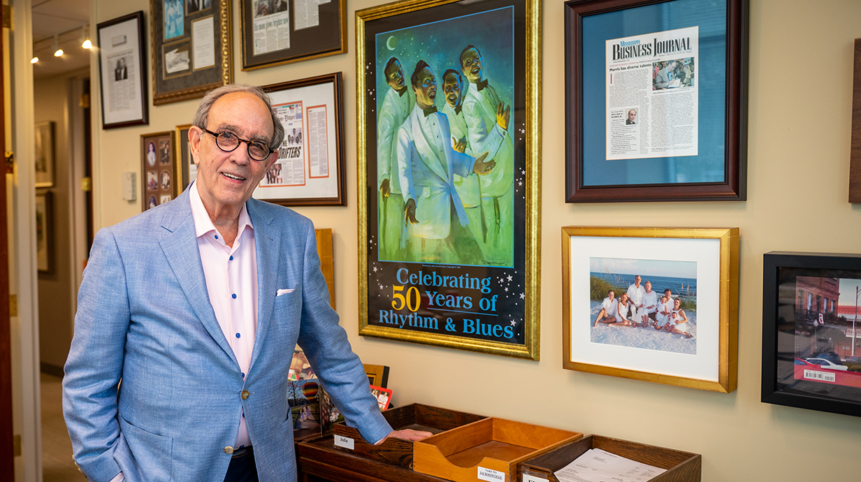 A man stands by a wall filled with framed artwork and news clippings.