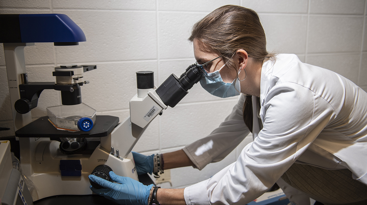 A woman wearing a lab coat looks through a microscope in a laboratory.