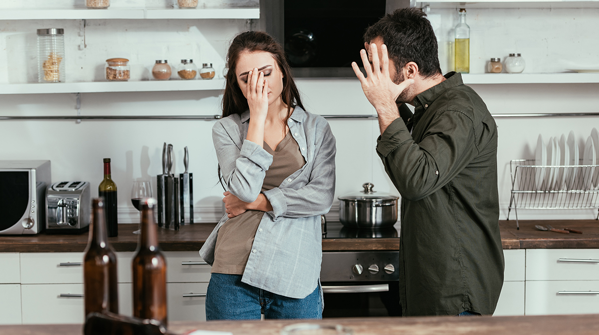 A man and woman argue in a kitchen with beer bottles sitting on the counter.