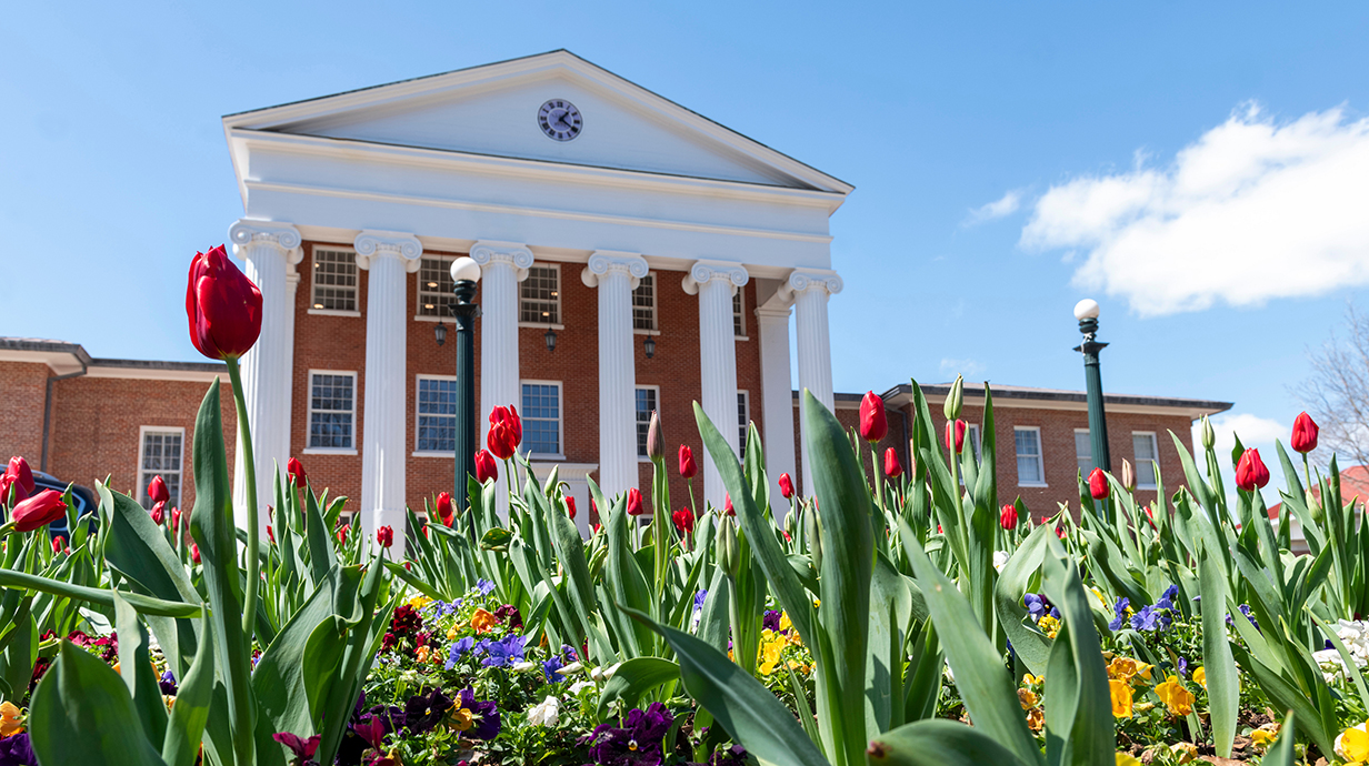 Colorful flowers bloom in front of a large red brick building with white columns lining the facade.