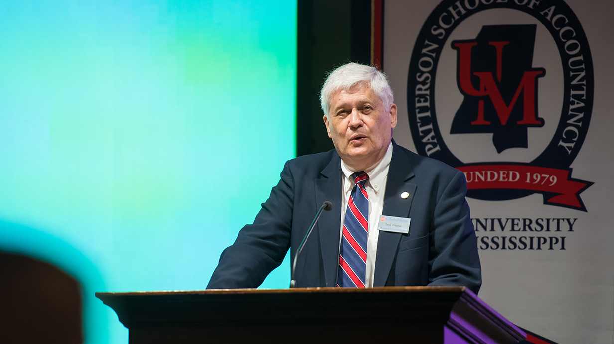 An older man wearing a suit speaks from a podium in front of a projection screen.