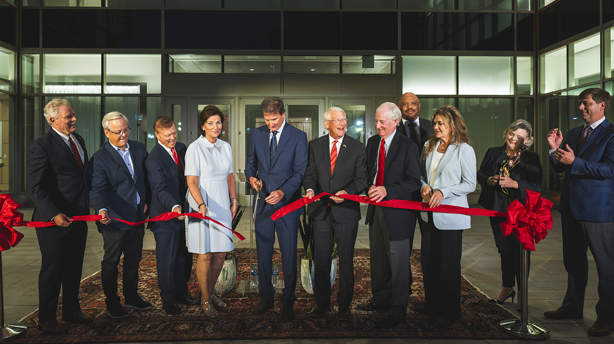 A large group of people watch while a man in the center cuts a red ribbon in front of a large building.