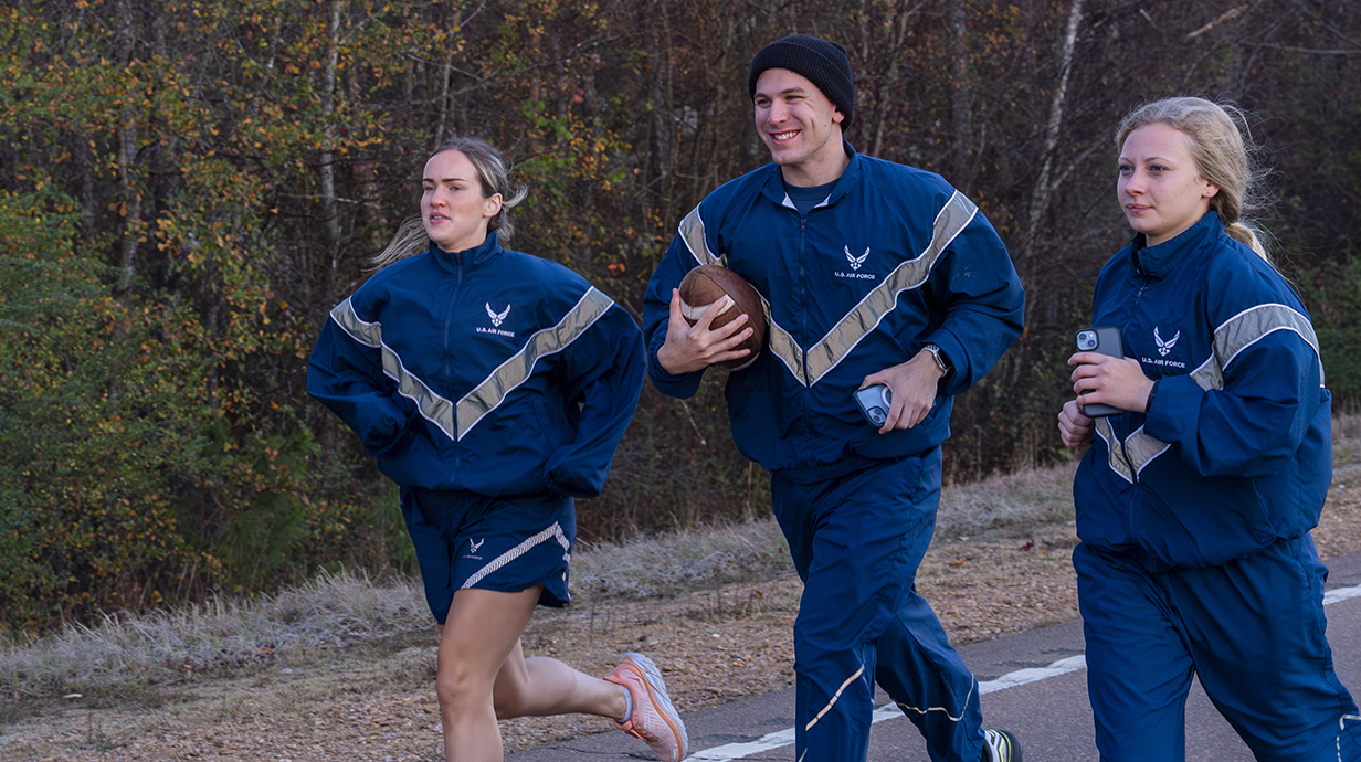 Two young women and a young man carrying a football -- all wearing matching U.S. Air Force running suits -- run along a highway.