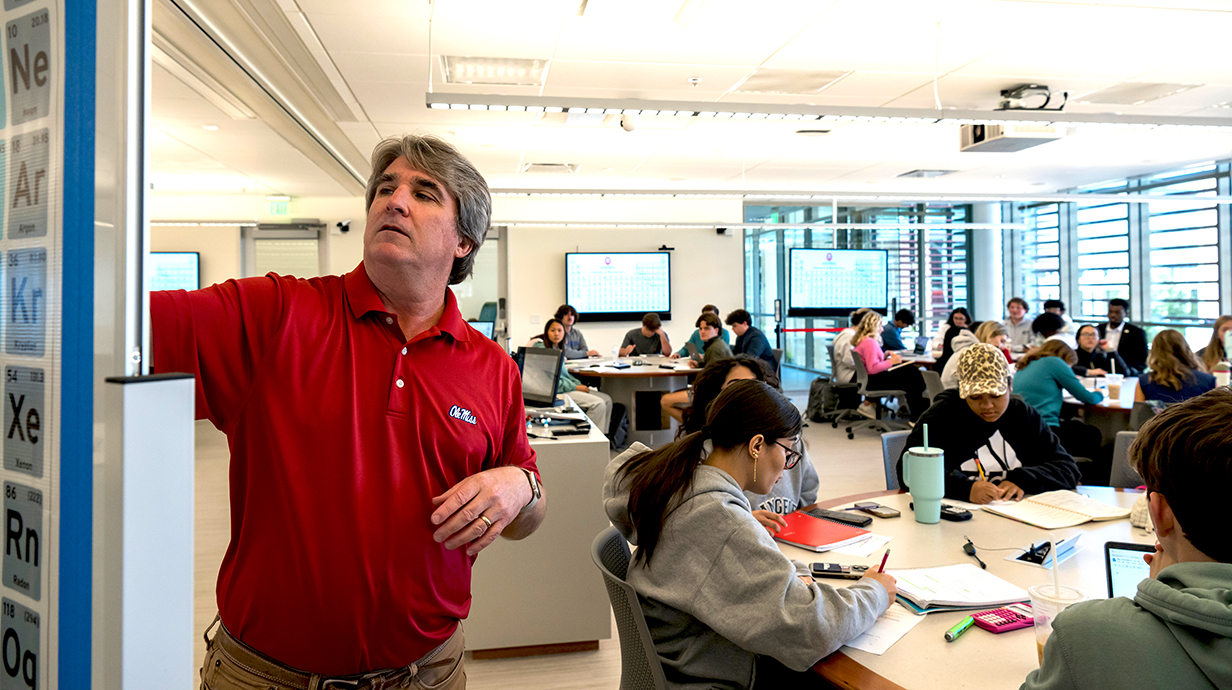 A teacher writes on a white board while a classroom of students behind him watch.