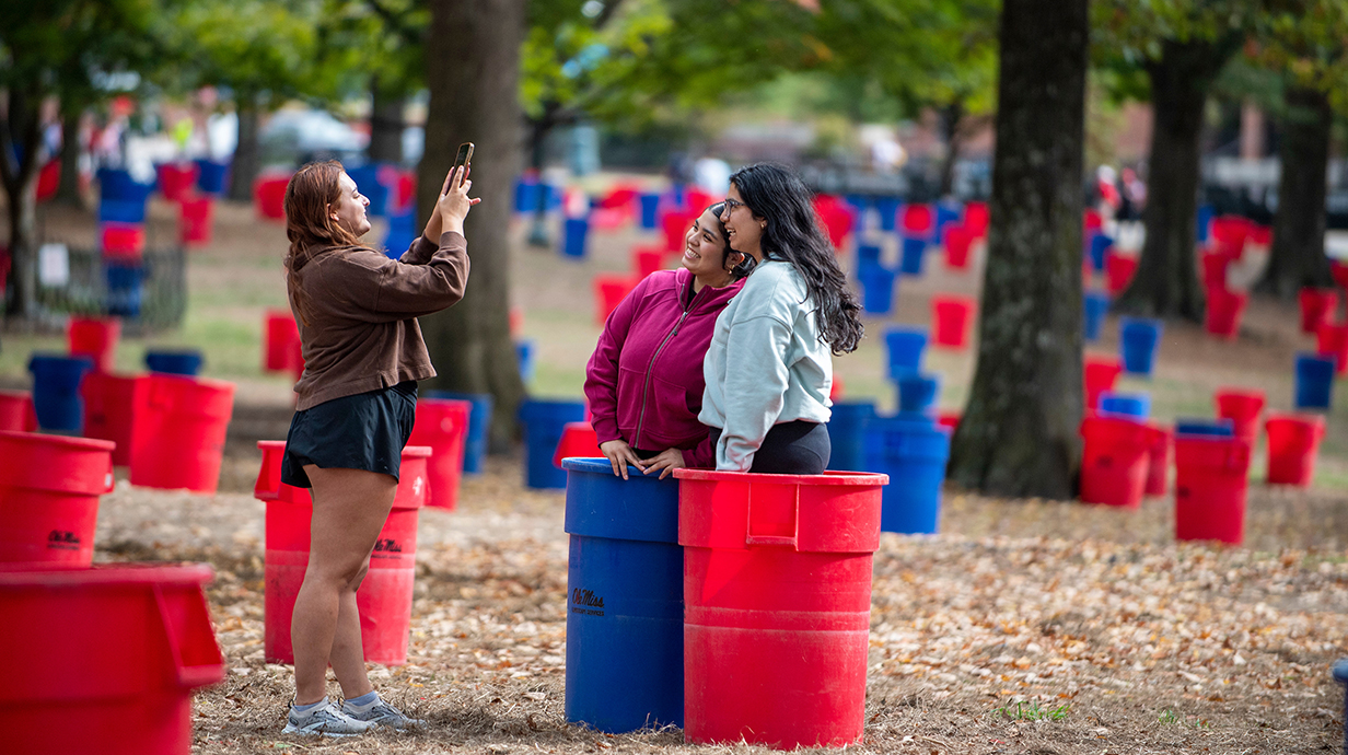 A young woman takes a photo of two other young women in a tree-lined park dotted with red and blue trash cans.