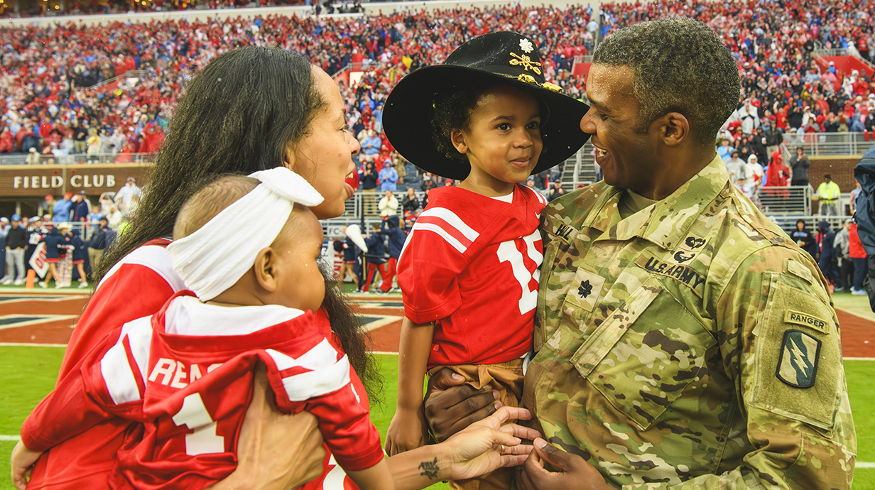 A man wearing military fatigues holds a young boy wearing a hat while standing on a football field with a woman holding a toddler.