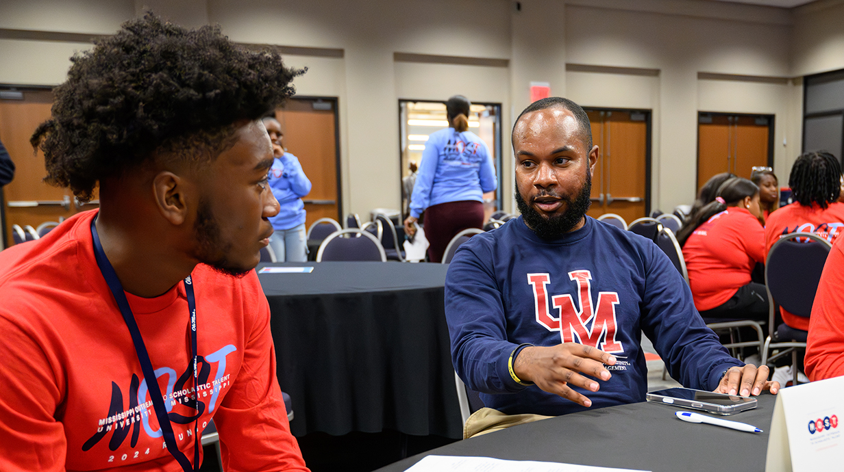 Two young men talk while sitting at a table in a banquet hall.