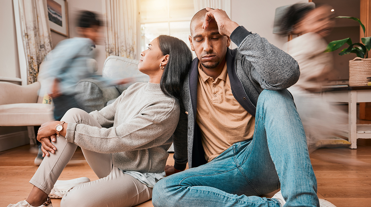 Two stressed-out adults sit with heads bowed while children run by in a blur.