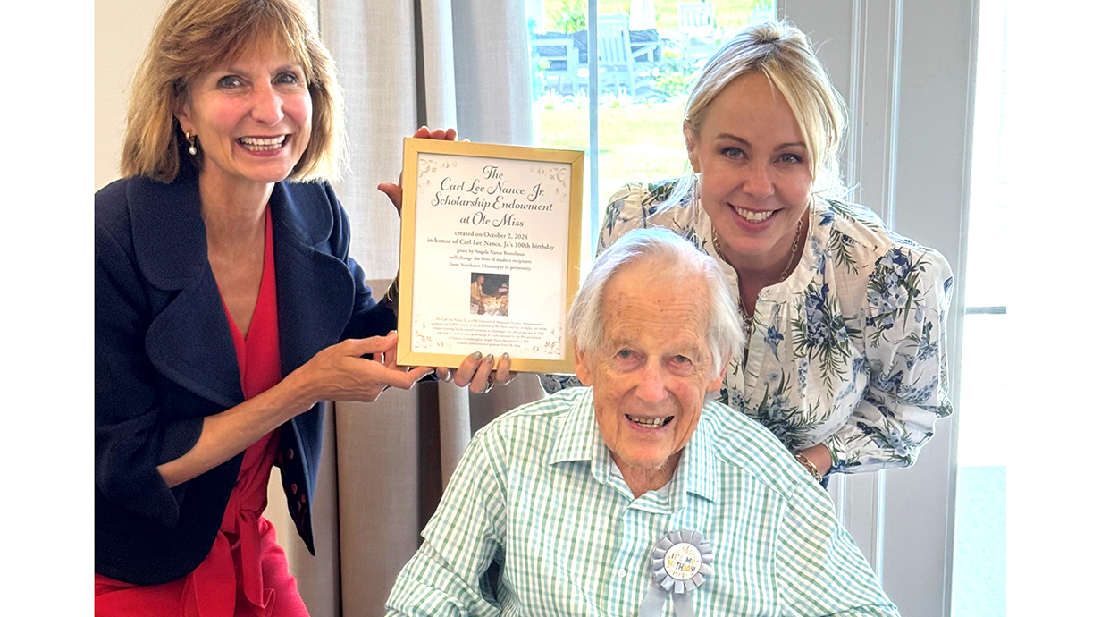 Two women holding a framed certificate stand behind a seated older man.