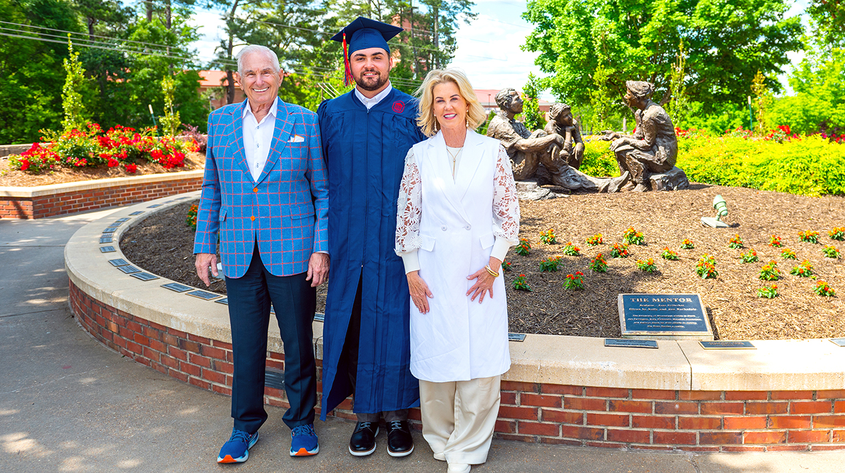 An older couple and a young man wearing graduation robes stand in front of a statue in an outdoor rose garden.