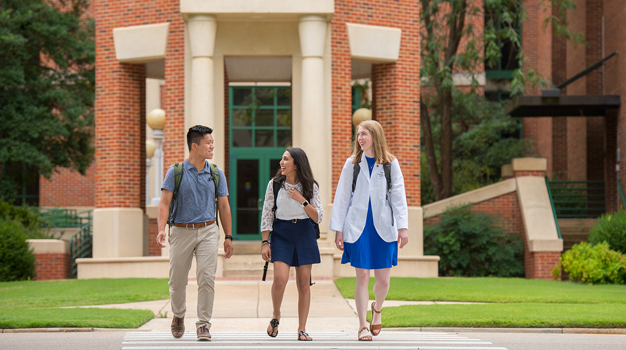 Two young women -- one wearing a white pharmacist coat -- and a young man cross a street in front of a large red brick building with stone columns.