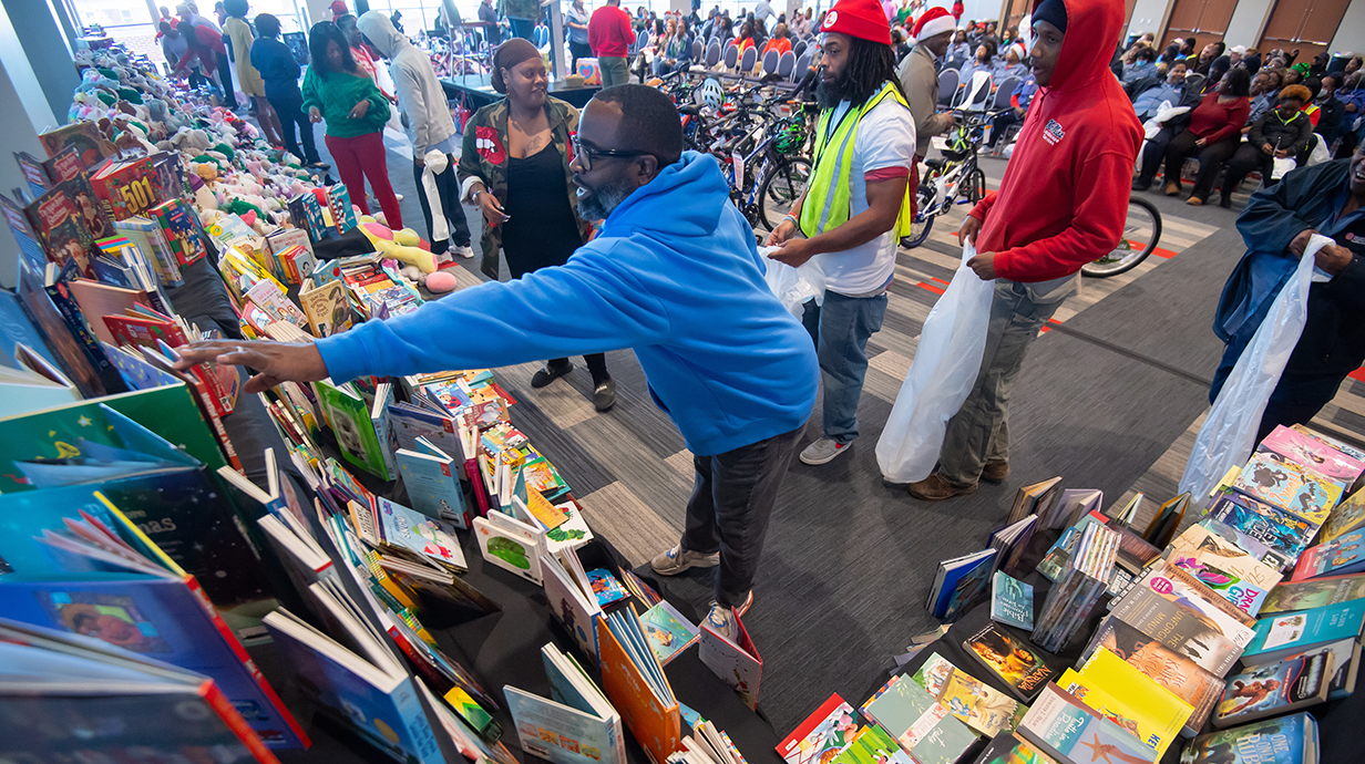 A man chooses a book from a large selection in a ballroom while two men holding bags wait their turn.