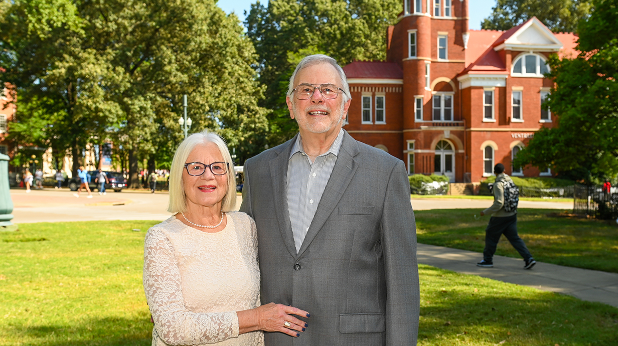 An older woman and man stand in a park in front of a red brick building.