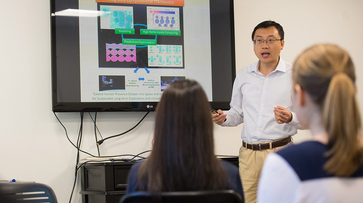 A man lectures in front of a projection screen in a classroom.