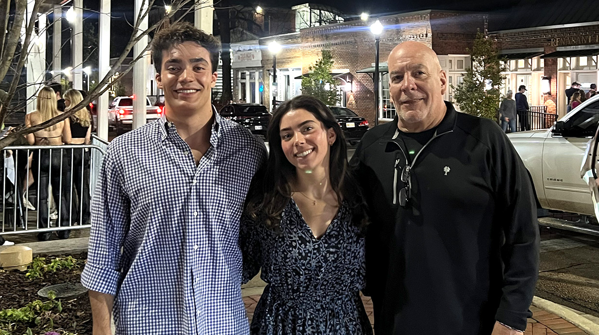 A man stands with a young man and woman on a street corner at night as cars drive behind them.