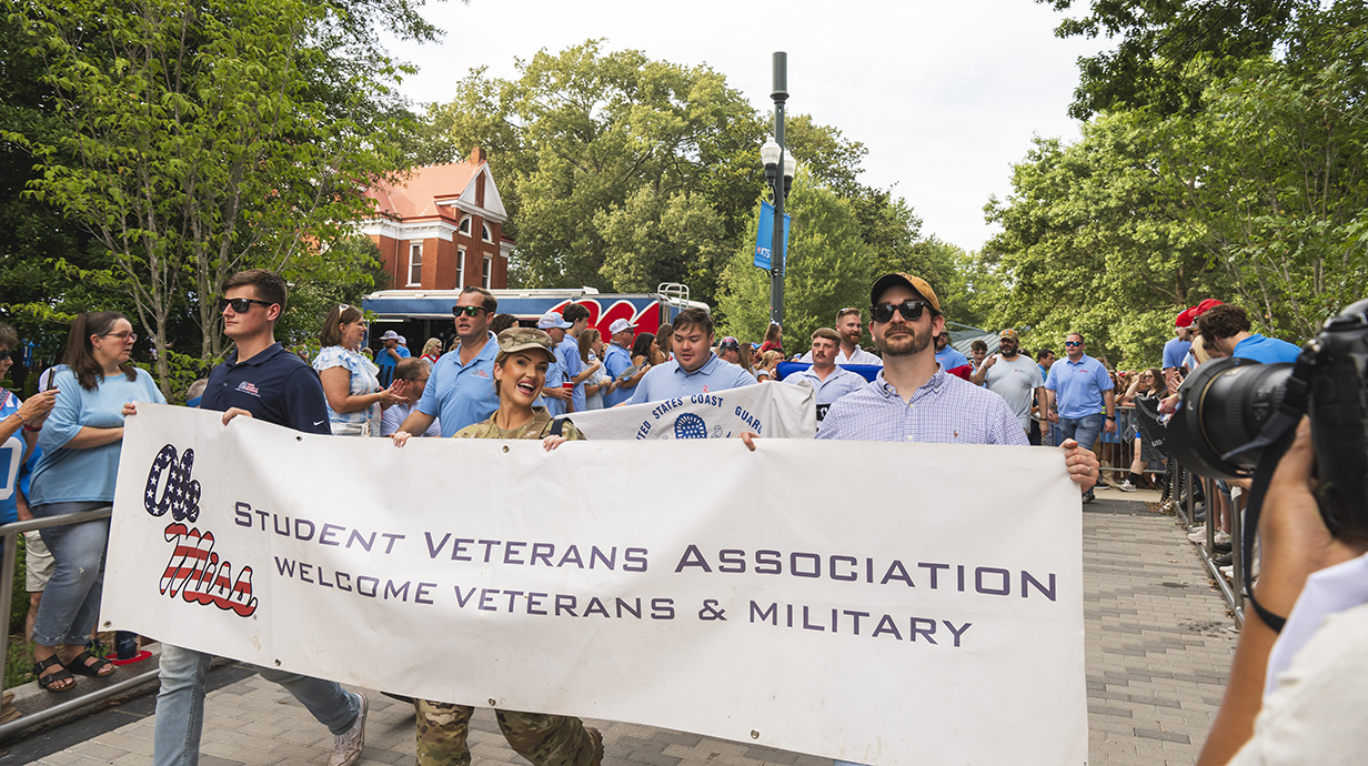 A group of men and a woman walk along a brick walkway carrying a banner that reads 'Ole Miss Student Veterans Association.'