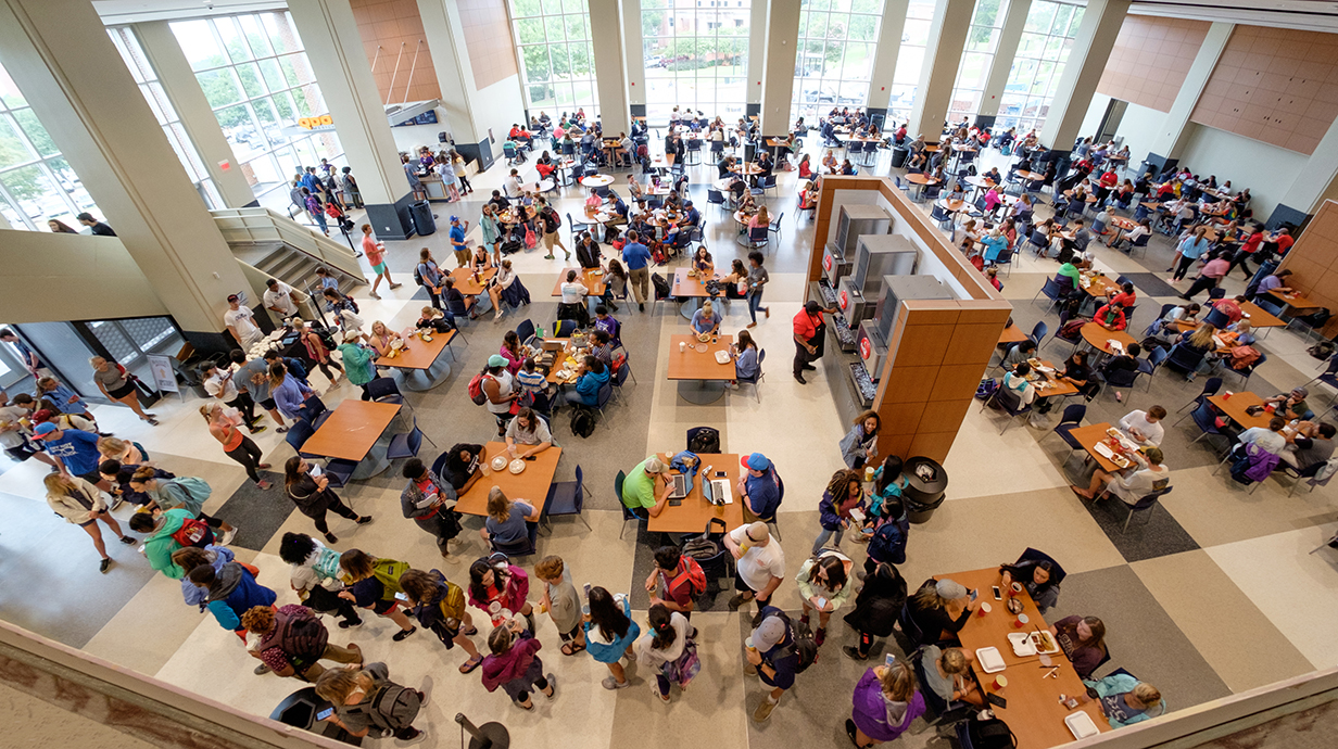 A wide shot of a food court from overhead includes hundreds of people sitting at tables and milling about.