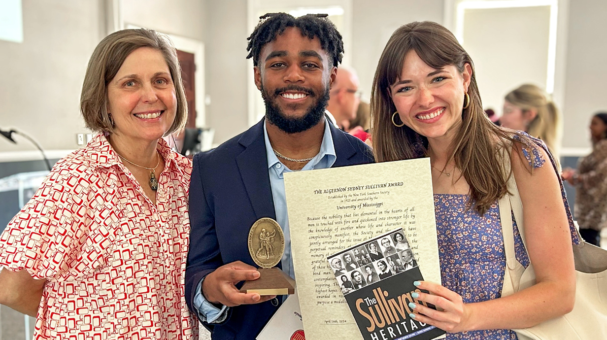 Two women pose with a young man holding a small award trophy and a certificate.