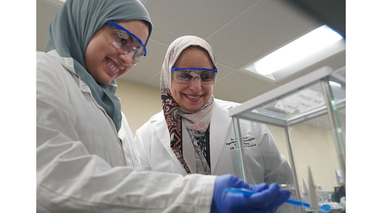 Two women wearing head wraps and safety glasses work in a laboratory.