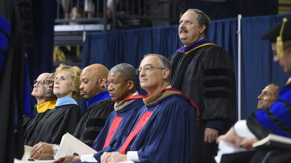 A man wearing graduation robes stands during a ceremony amid a crowd of robed people.