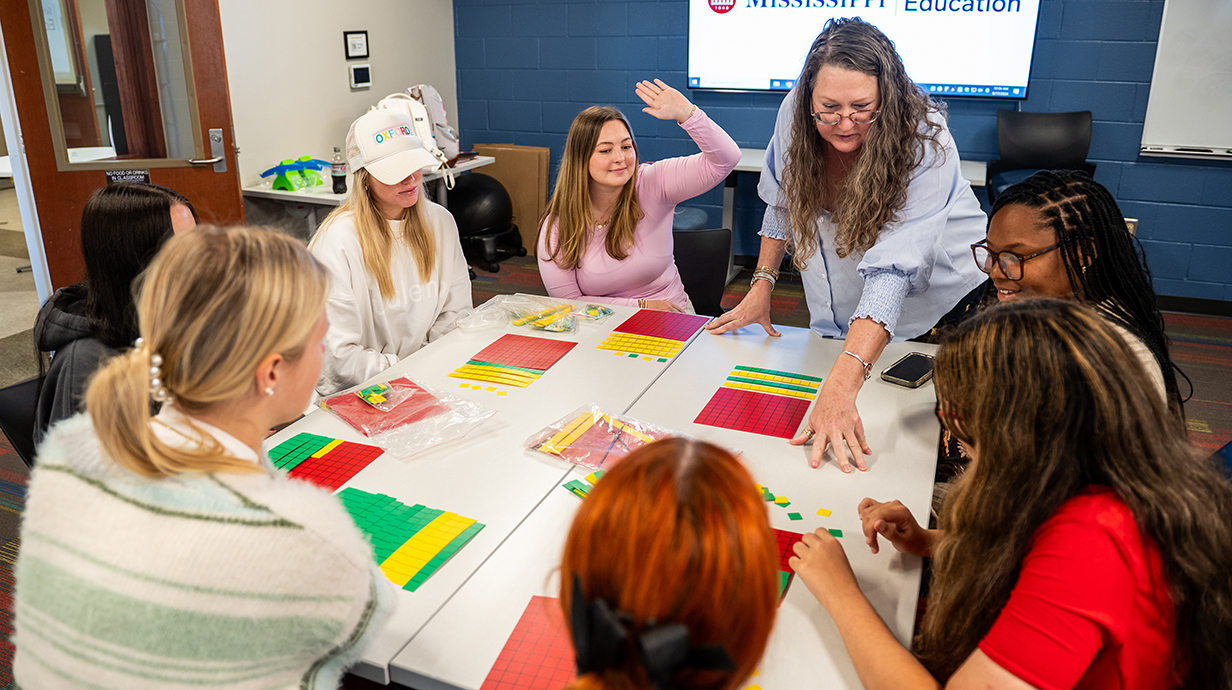 A teacher standing at a table with students seated all around points to colored tiles on the tabletop.