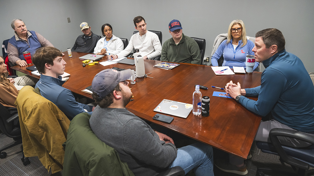 A group of people sitting around a conference table listen as a man sitting at the right end speaks.