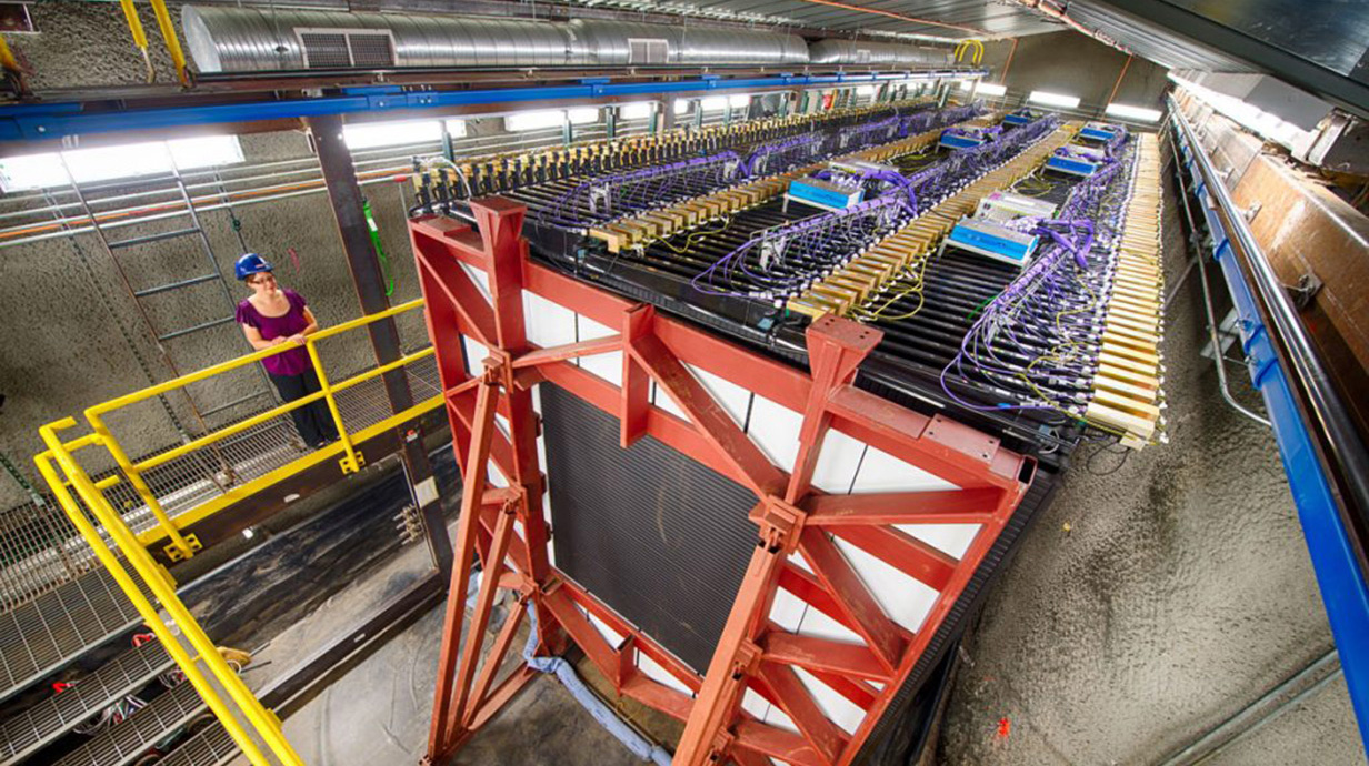 A woman wearing a blue hard hat stands on a platform overlooking a giant machine in a large building.