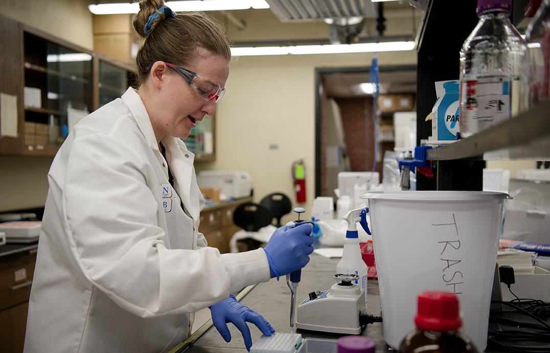 A woman wearing lab gear uses a pipette to transfer liquid to a tray in a laboratory.