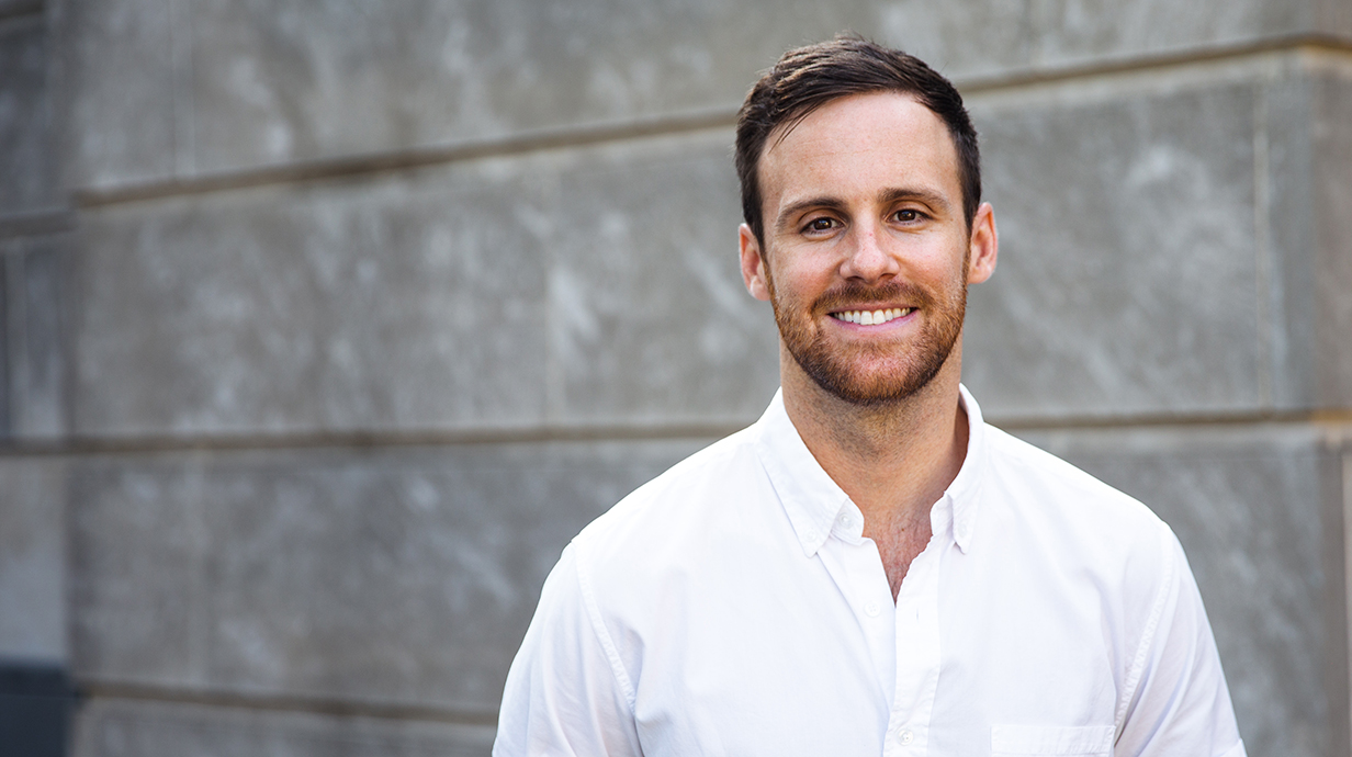 Outdoor portrait of a man wearing a white shirt standing in front of a gray block wall.