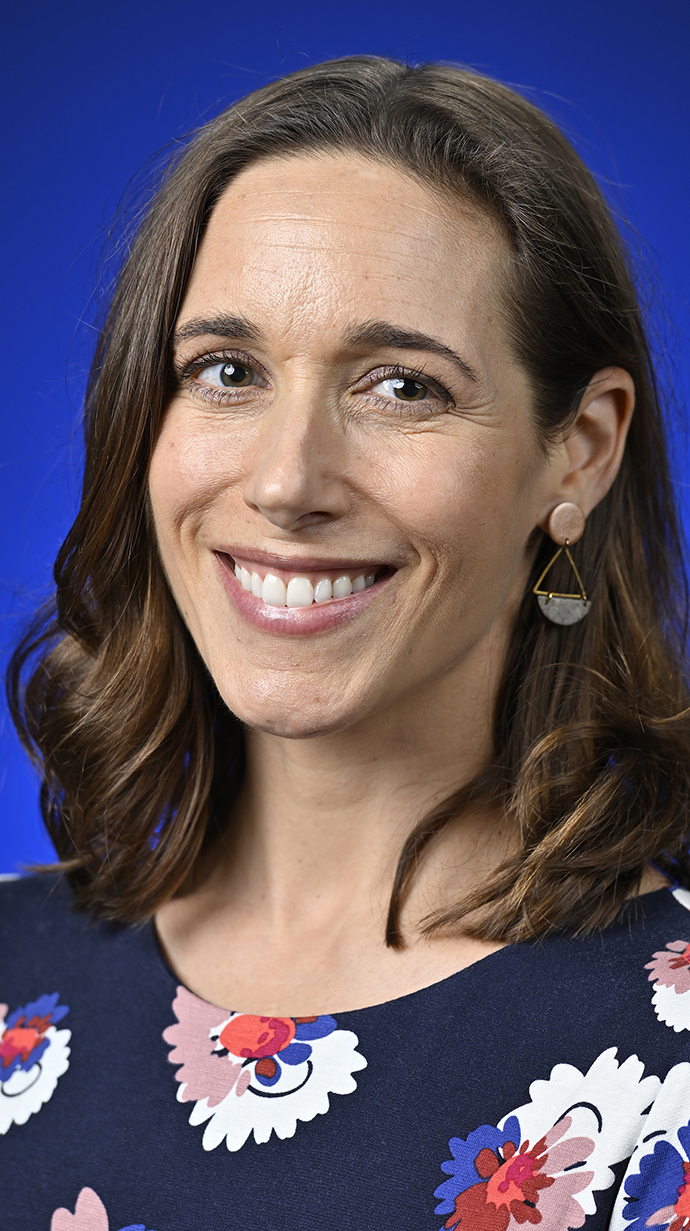 Headshot of a woman wearing a dark blue top decorated with colordul flowers.