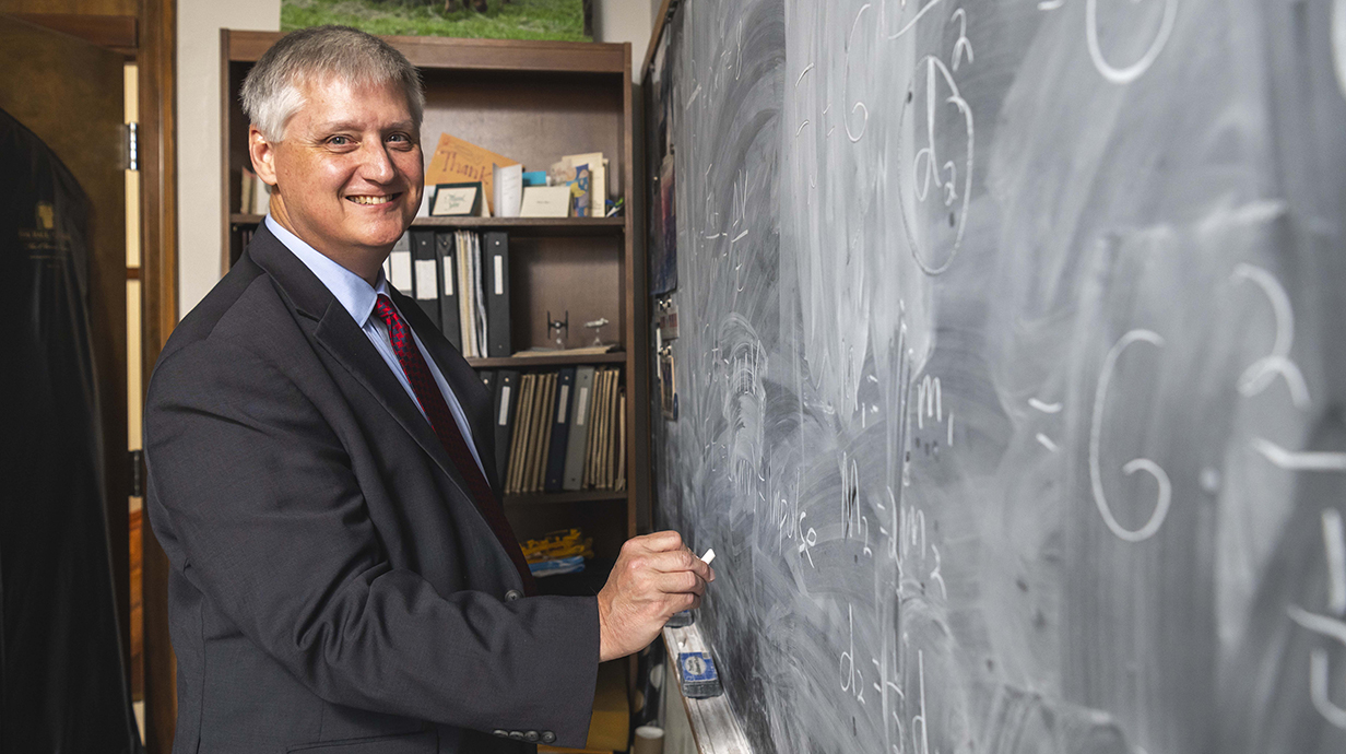A man wearing a suit works at a blackboard filled with equations.