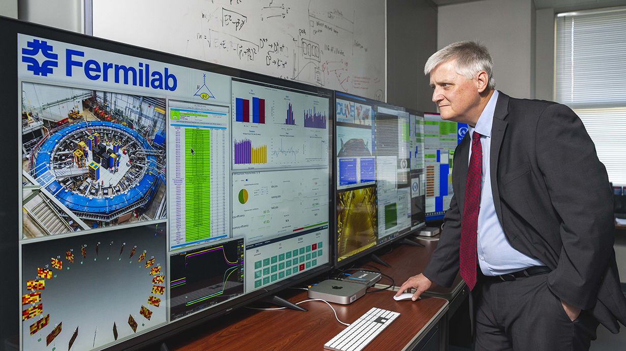 A man stands as he works with a computer attached to a large video monitor.