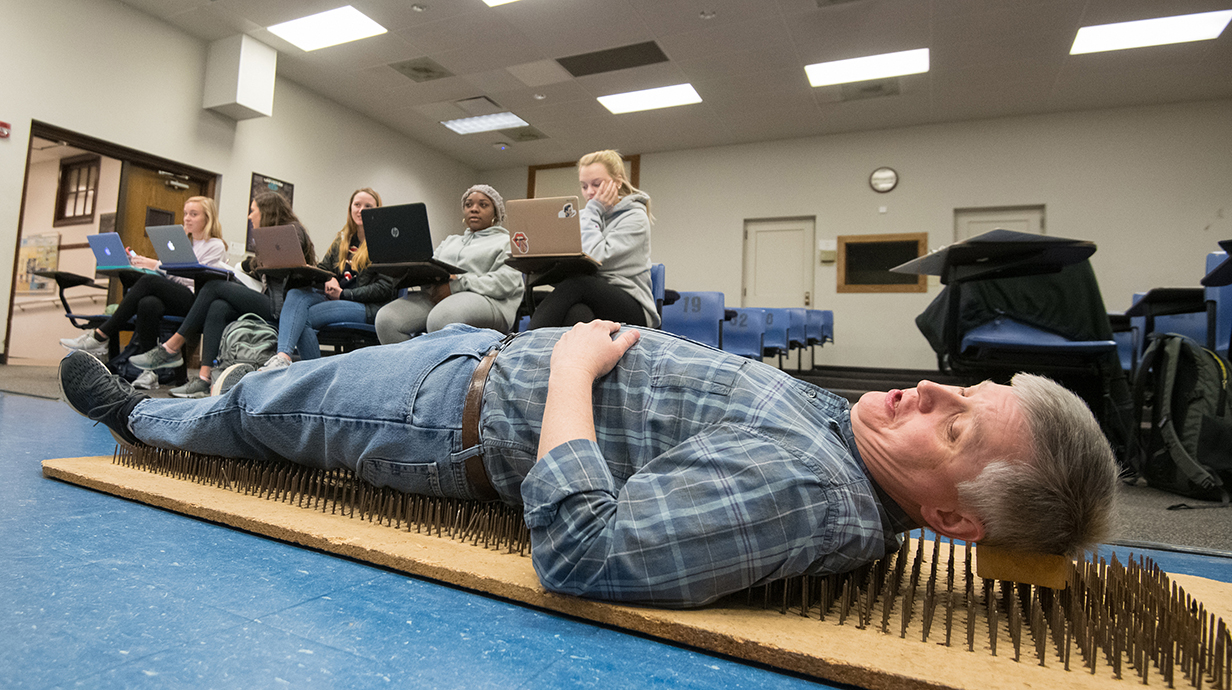 A man lies on a bed of nails in a classroom in front of a group of students.