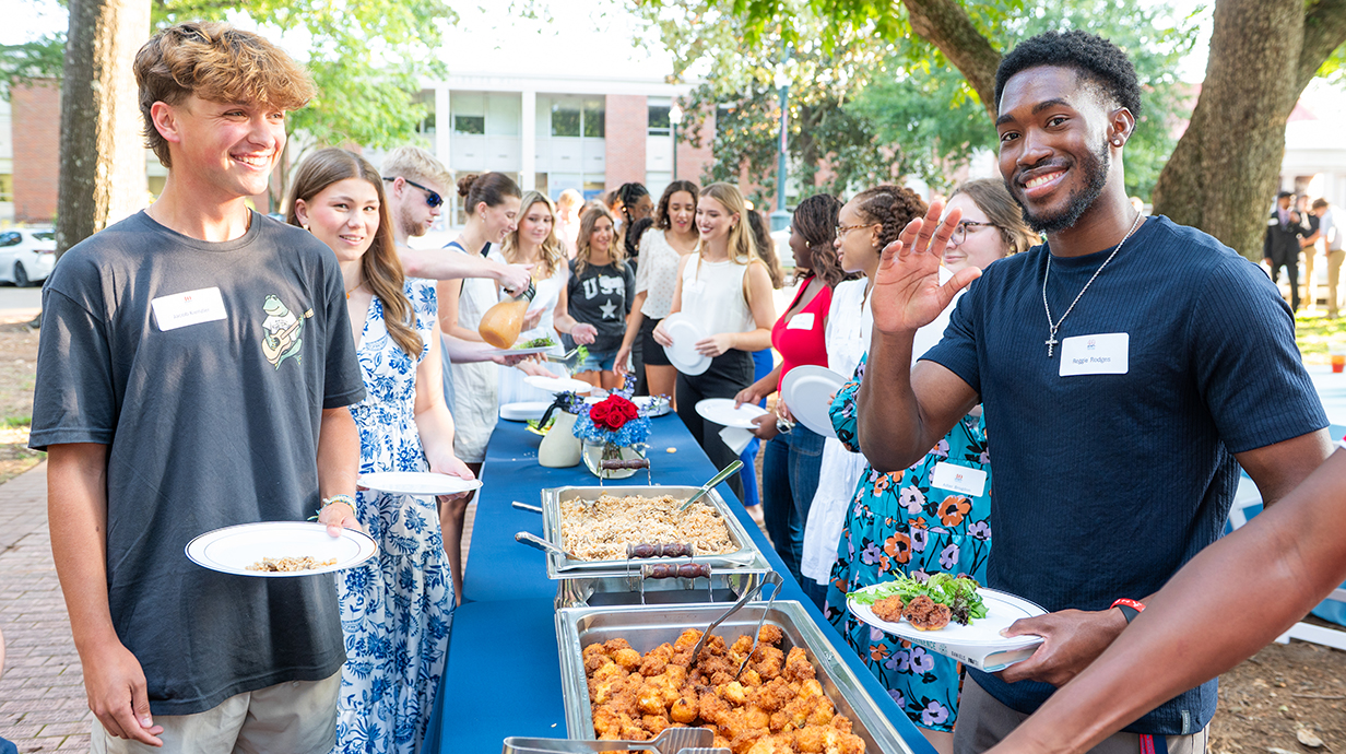 A group of young people fill their plates at an outdoor buffet in a park.