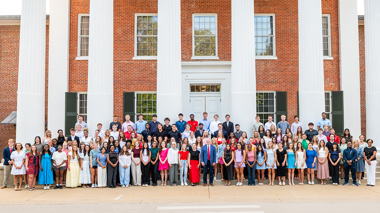 A large group of people stand on the steps of a three-story brick building with white columns.