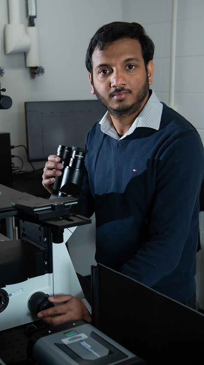 A man wearing a blue sweater sits at a microscope in a lab.