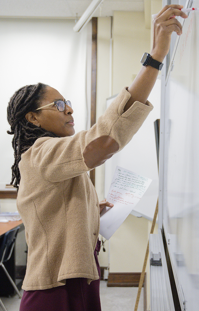 A woman reaches above her head to write on a whiteboard in a classroom.