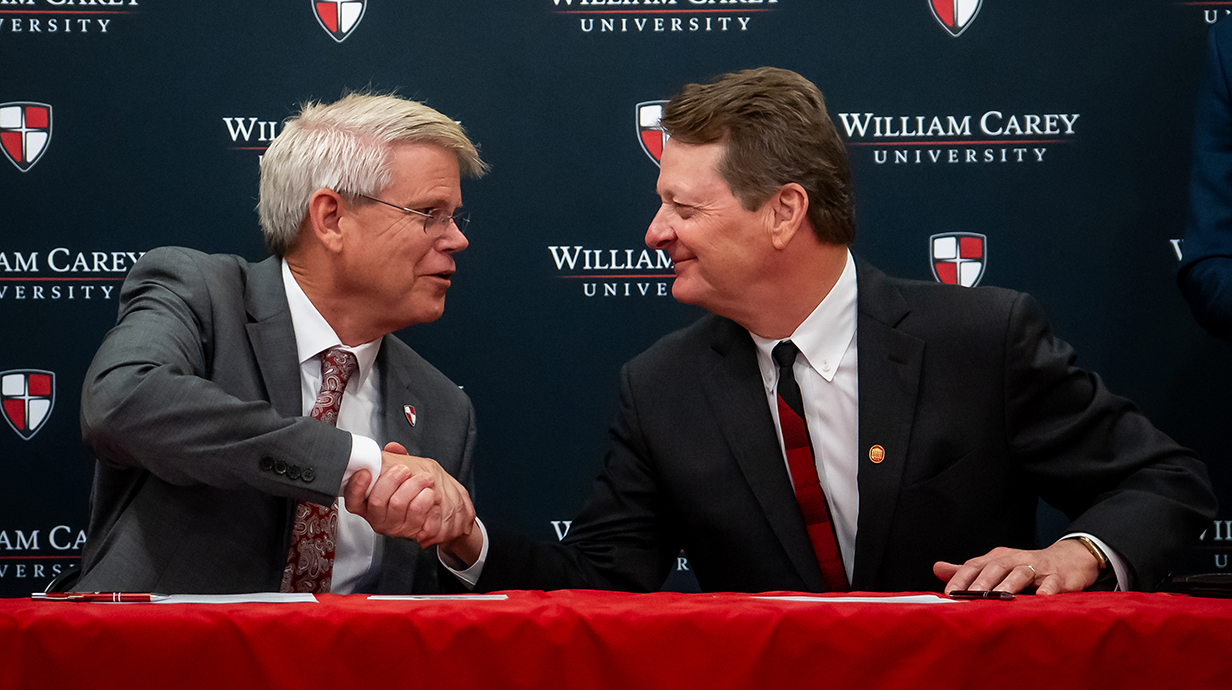 Two men shake hands while seated next to each other at a table.