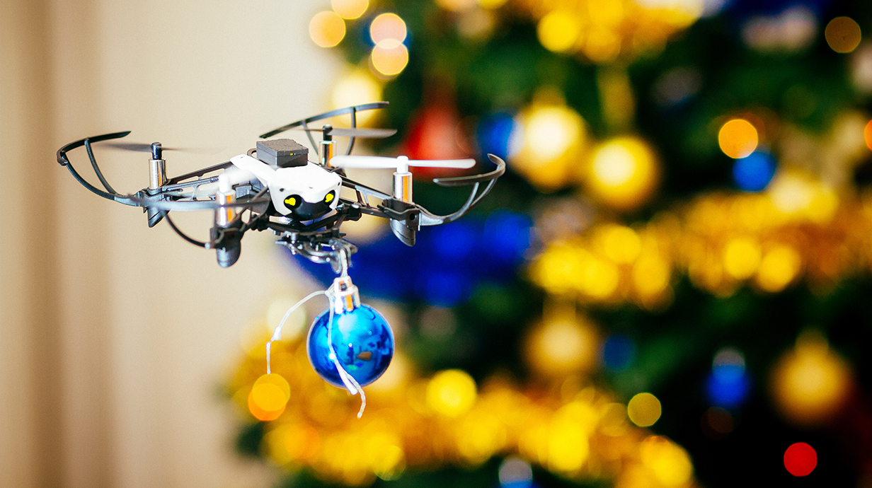 A small drone holding a blue Christmas ornament flies in front of a Christmas tree.