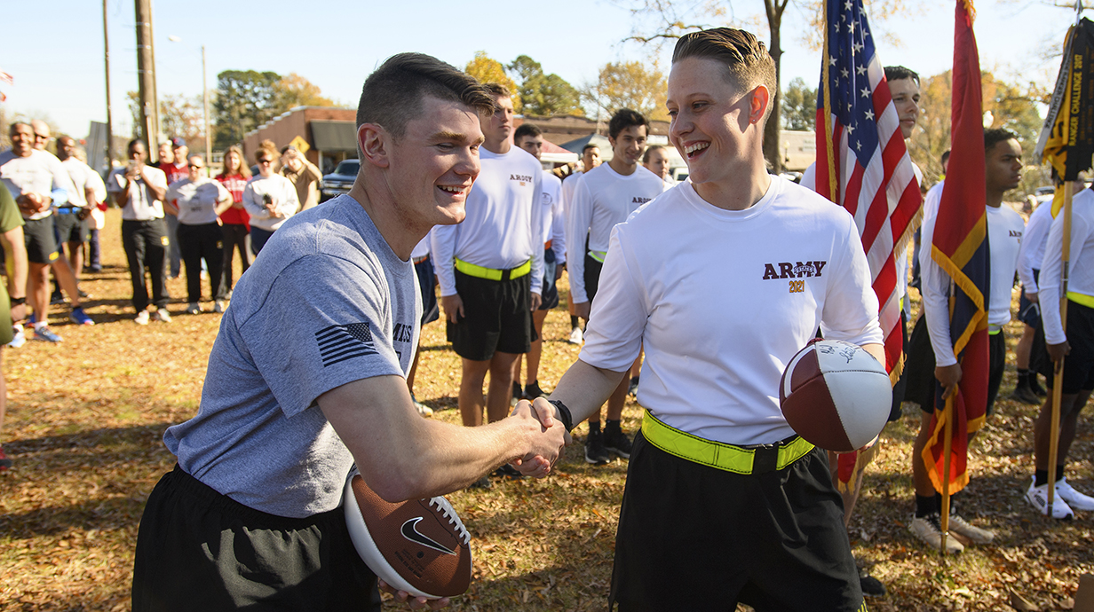 A young man and a young woman, both holding footballs, shake hands in an outdoor park.