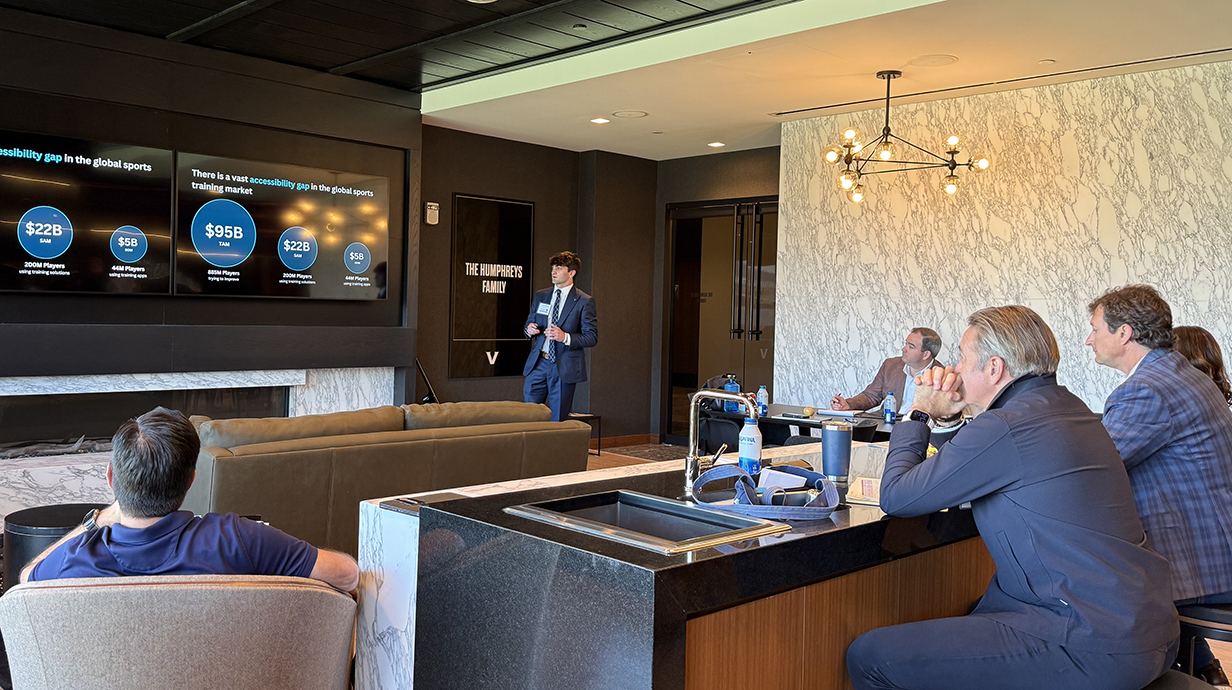 A young man talks in front of a projection screen as several seated men listen.