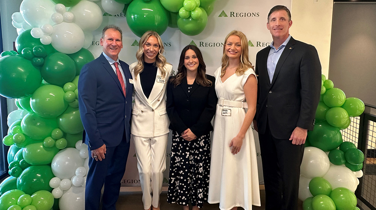 Two men and three young women stand in front of a backdrop covered in green and white balloons.