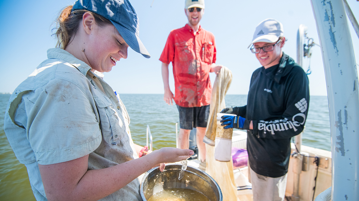 A young woman checks a water sample which standing on the deck of a boat as two young men watch.