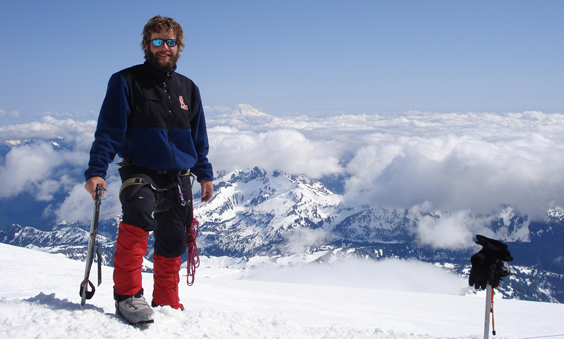 A bearded man wearing a blue coat and red ski pants stands atop a snow-covered mountain on a clear day.