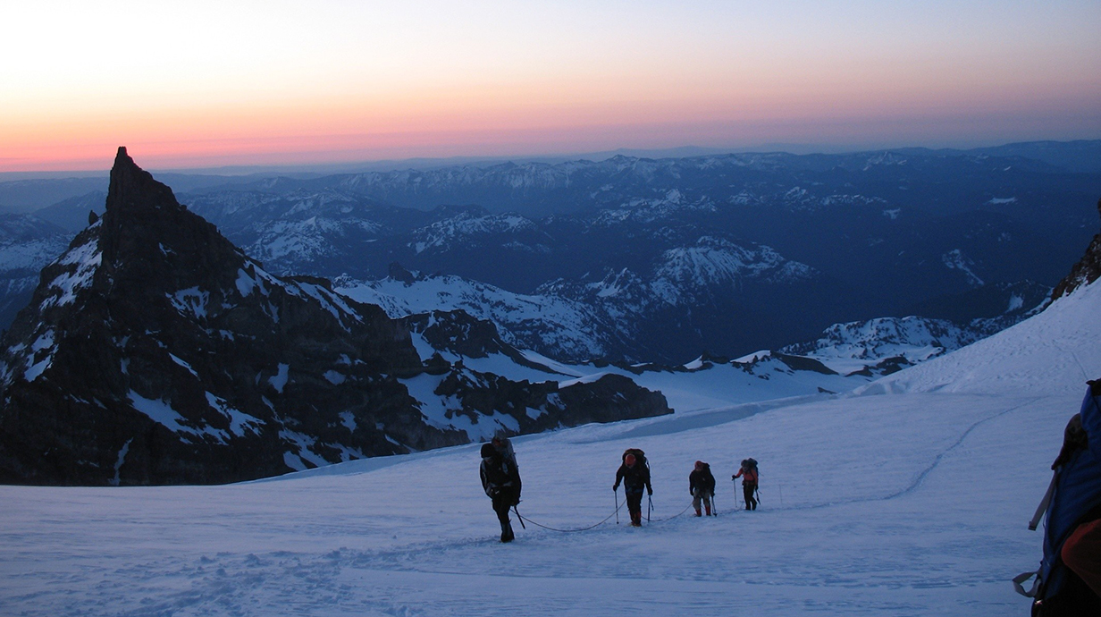 Four climbers work their way up a snowy slope of a mountain with a scenic valley in the background.