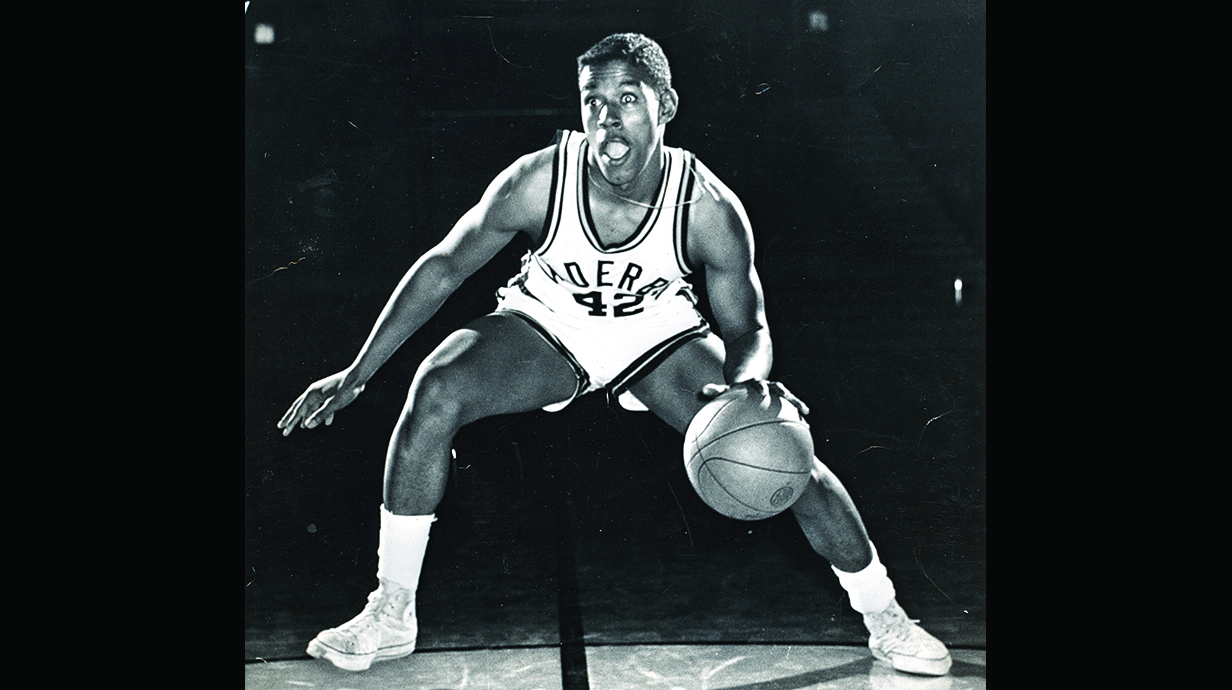 A black-and-white promotional photo of a basketball player dribbing a basketball on a court.