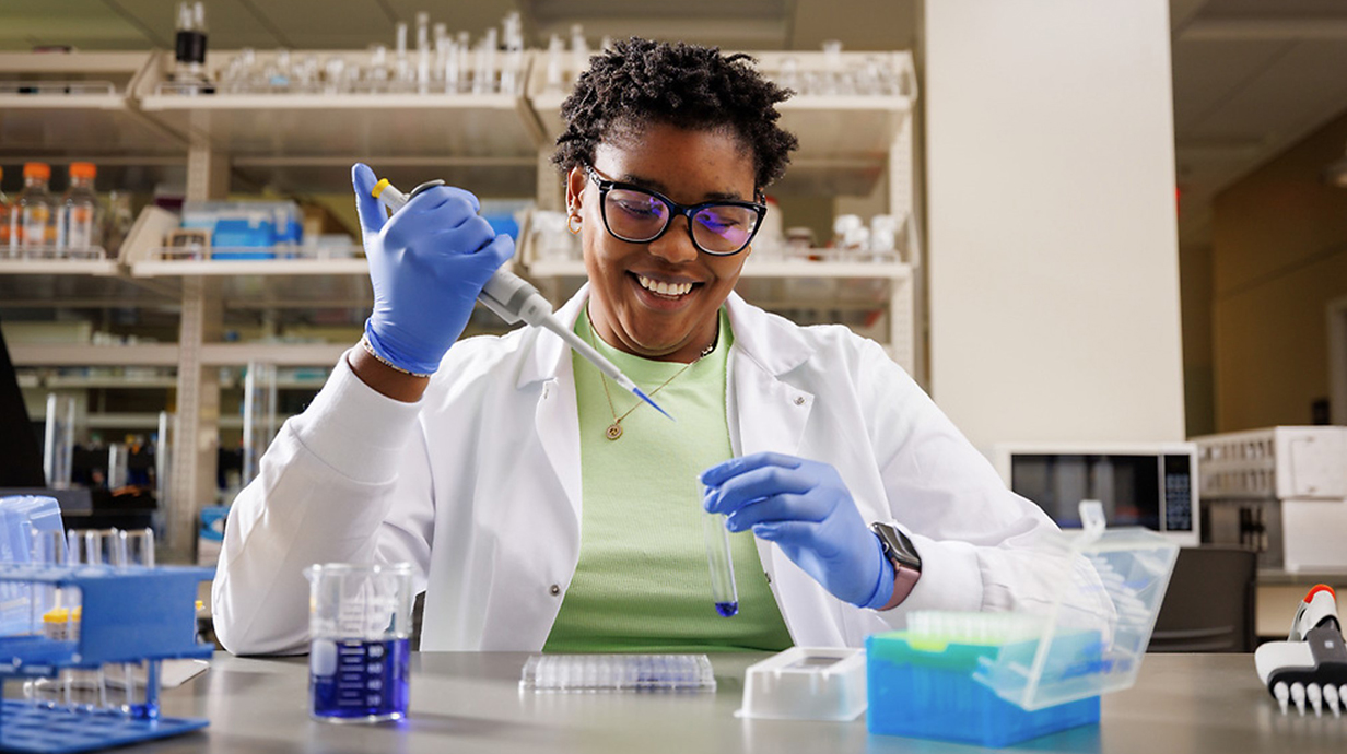A woman wearing a white lab coat and blue gloves uses a pipette to transfer blue liquid to a test tube in a laboratory.