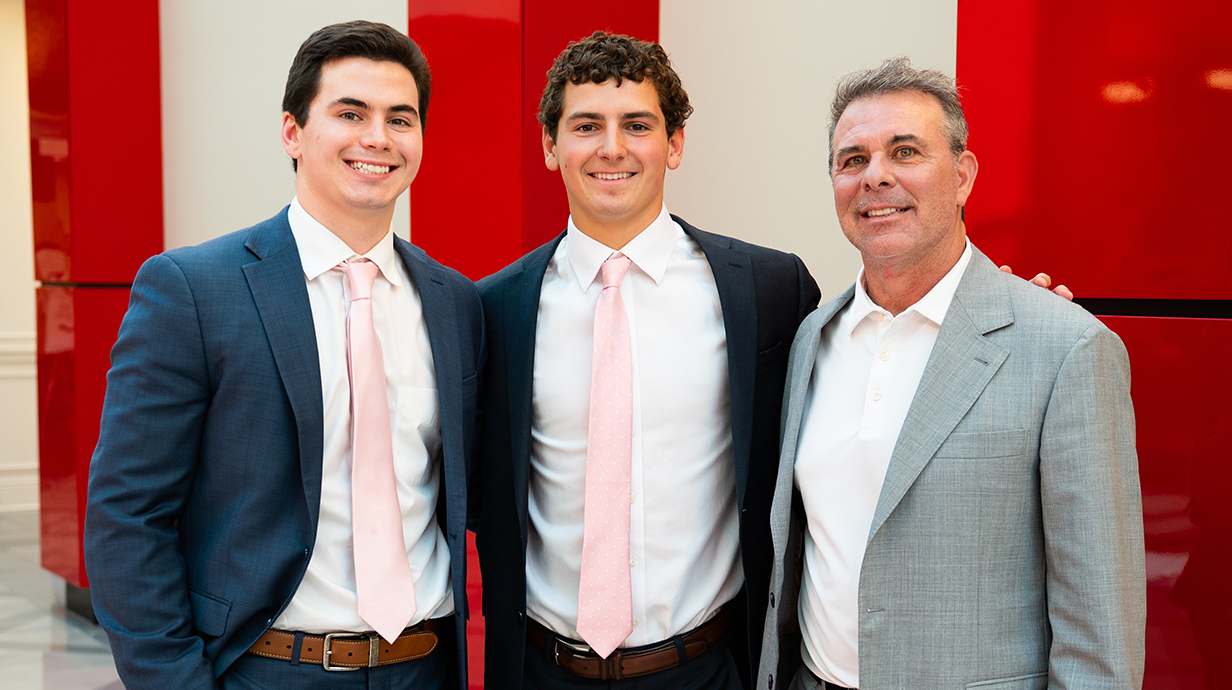 A man wearing a sport coat poses for a photo with two young men in suits in a hallway.