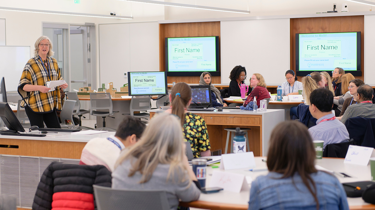 A woman talks to a classroom of people sitting around tables lined with computers.
