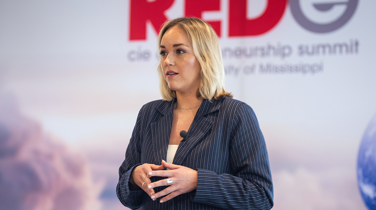 A young woman speaks at a conference in front of a colorful backdrop.